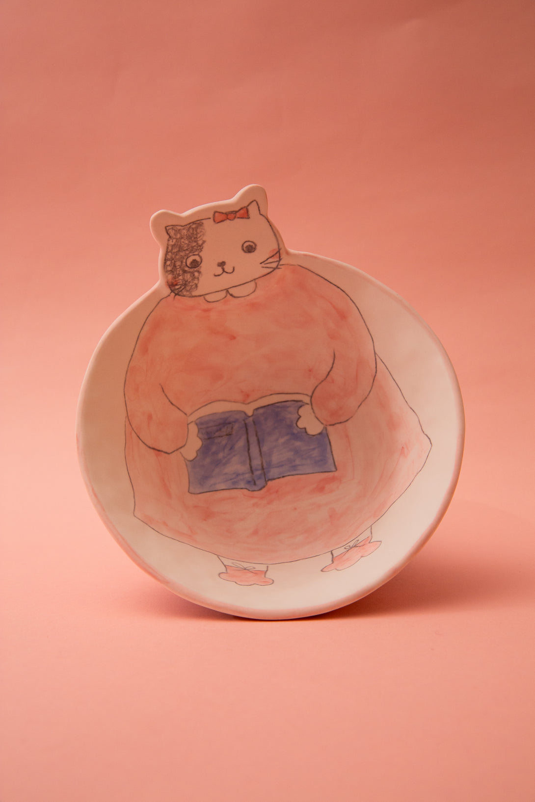 Ceramic bowl with a cartoon cat holding a book on a pink background