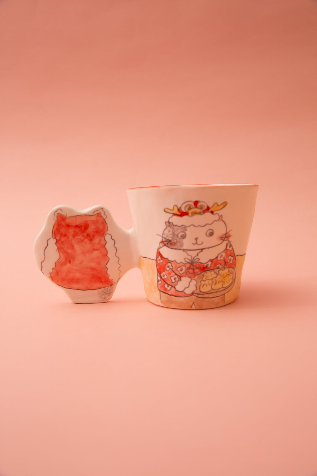 A ceramic mug with cat illustrations and a fun handle on a pink background