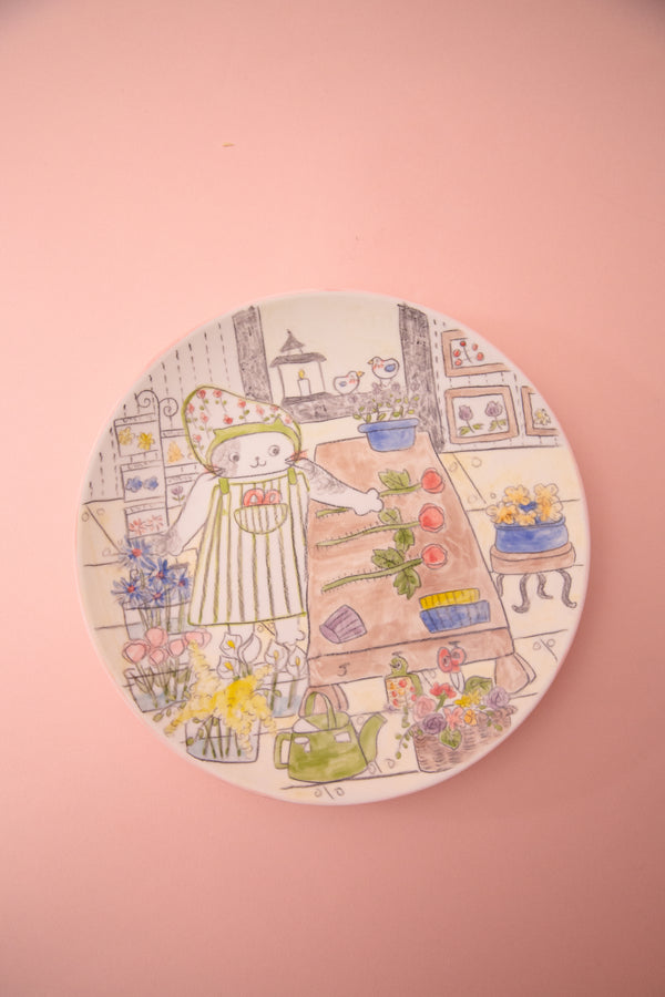 Circular plate with illustration of a cartoon cat in a flower shop with various items on a pink background