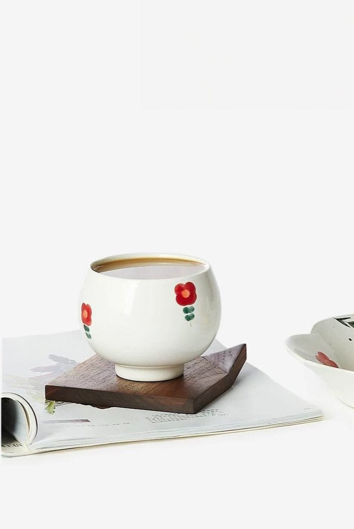 A white ceramic cup with red and green floral patterns on the side, displayed against a white background on a book and wooden coaster.