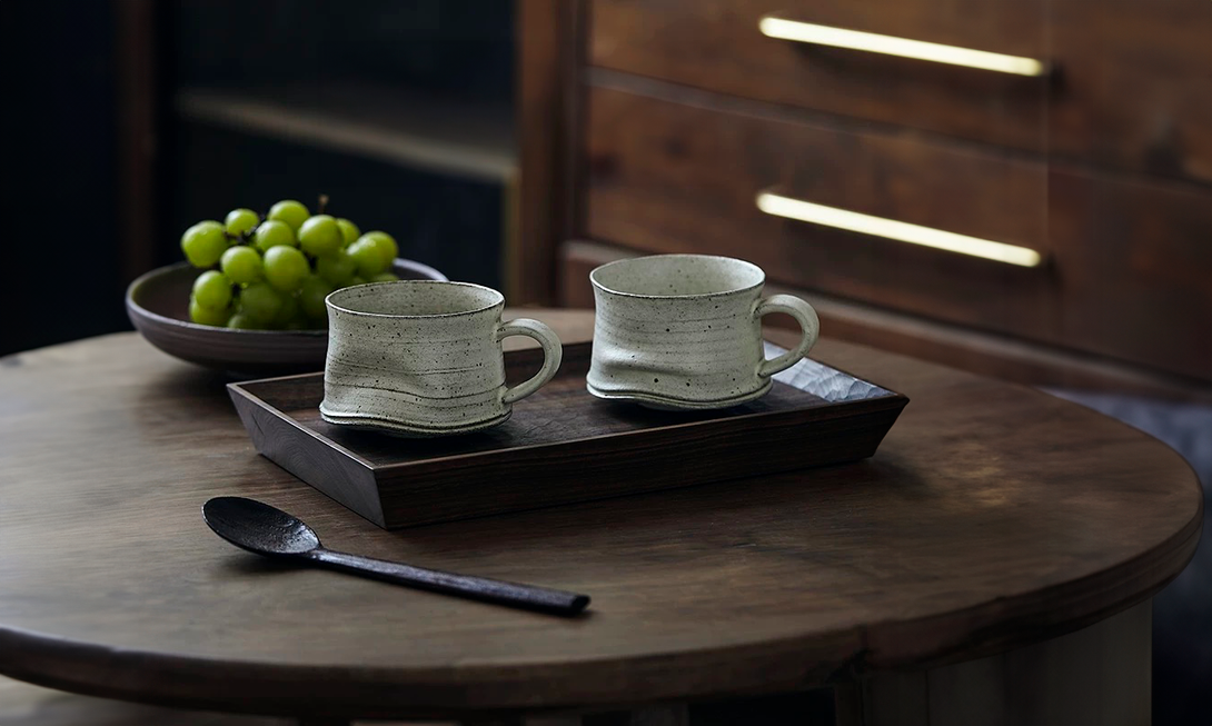Two ceramic mugs on a wooden tray with grapes and a spoon on a wooden table.