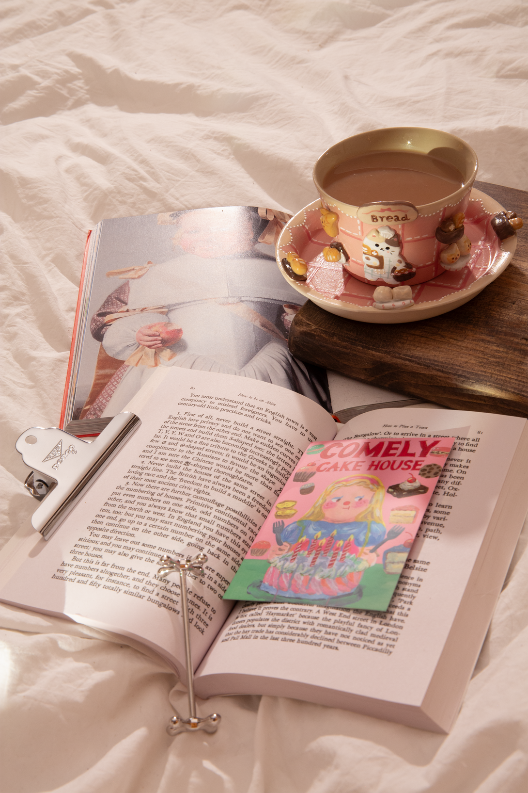 Open book with a bookmark, a cute pink cup holding coffee, and a magazine on a bed.