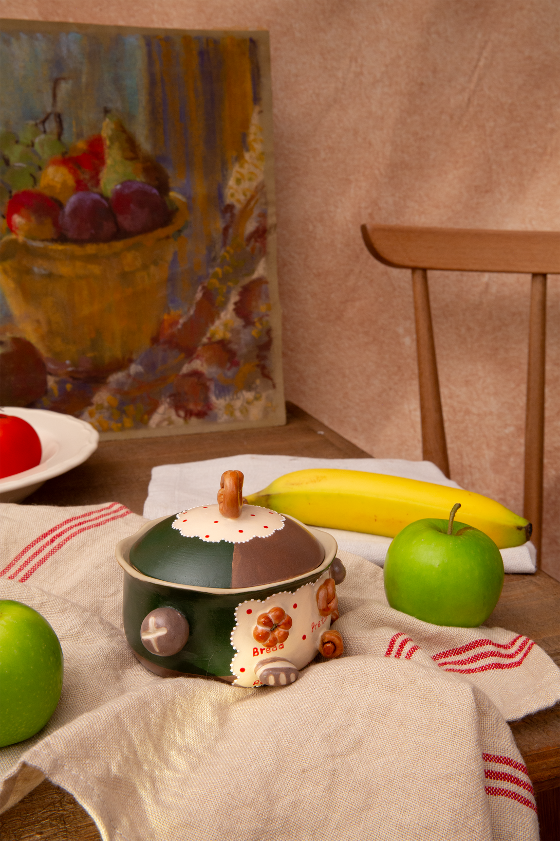 ceramic bowl with fruit on a tablecloth, with a painting of fruit in the background.