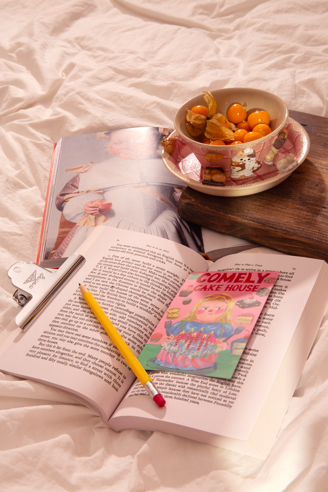 Open book with a pencil, colorful bookmark, and a bowl of fruit on a bed.