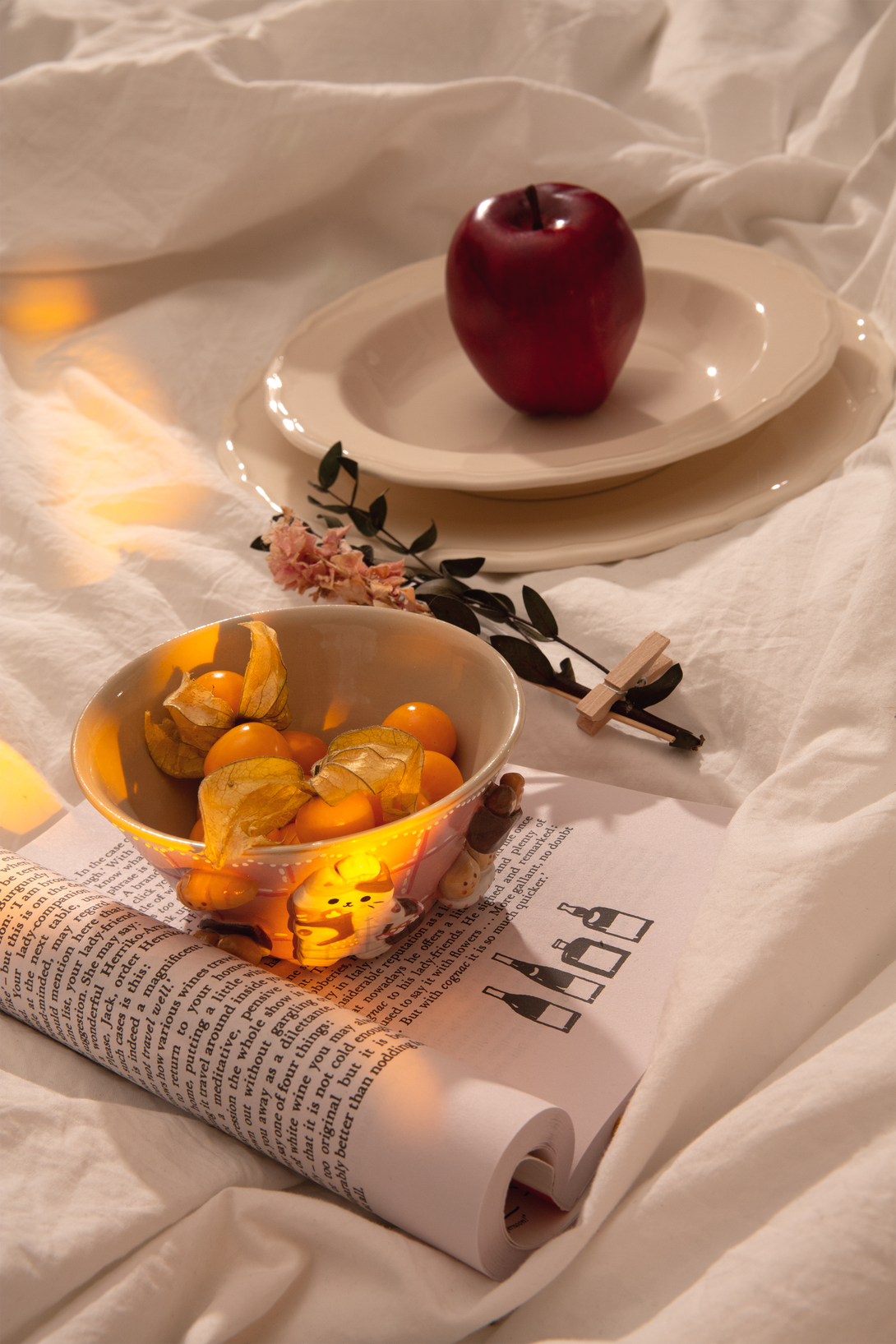 Apple on a white plate with a bowl of fruit and an open book on a white surface