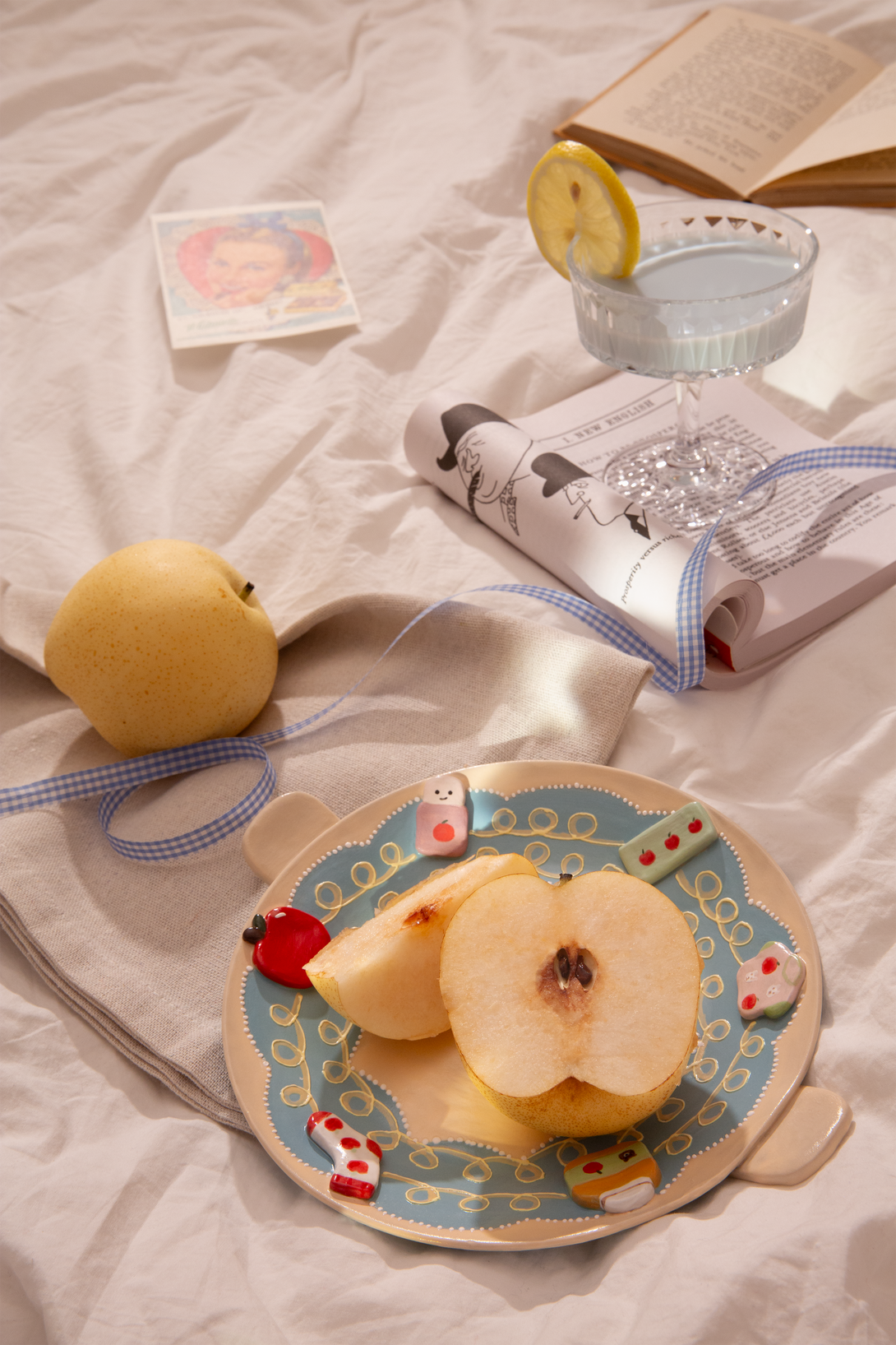 Sliced pear on a ceramic plate with handles, a glass of lemonade and a book on a light surface
