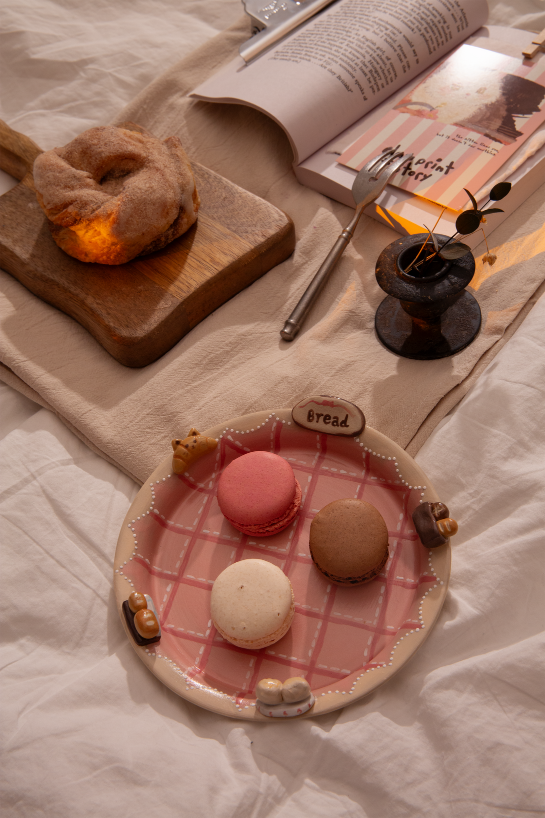 Plated macarons on a checkered pink plate with a bagel and book on a white surface