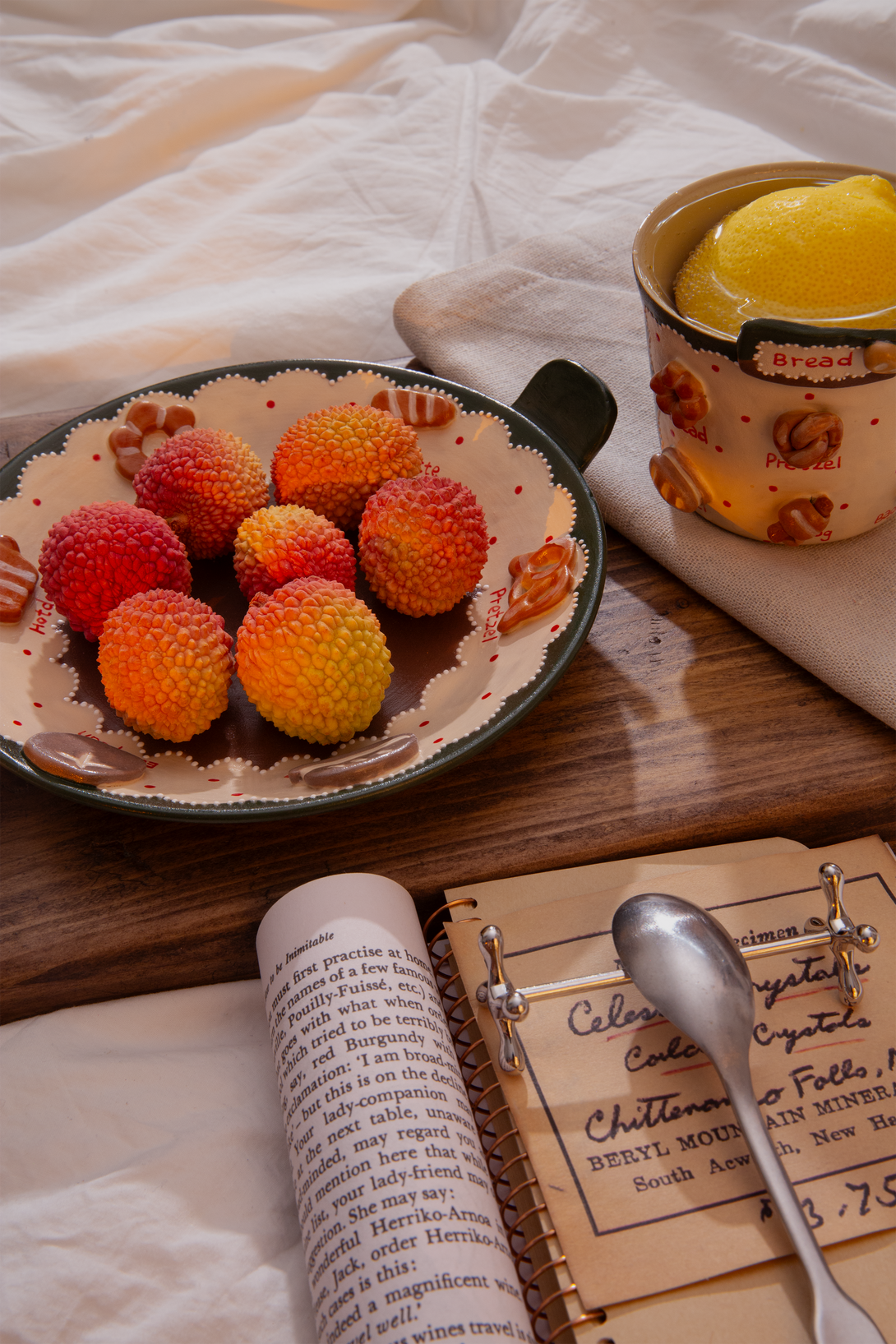 Plate of lychees, a mug with a yellow dessert, and an open book on a wooden surface.