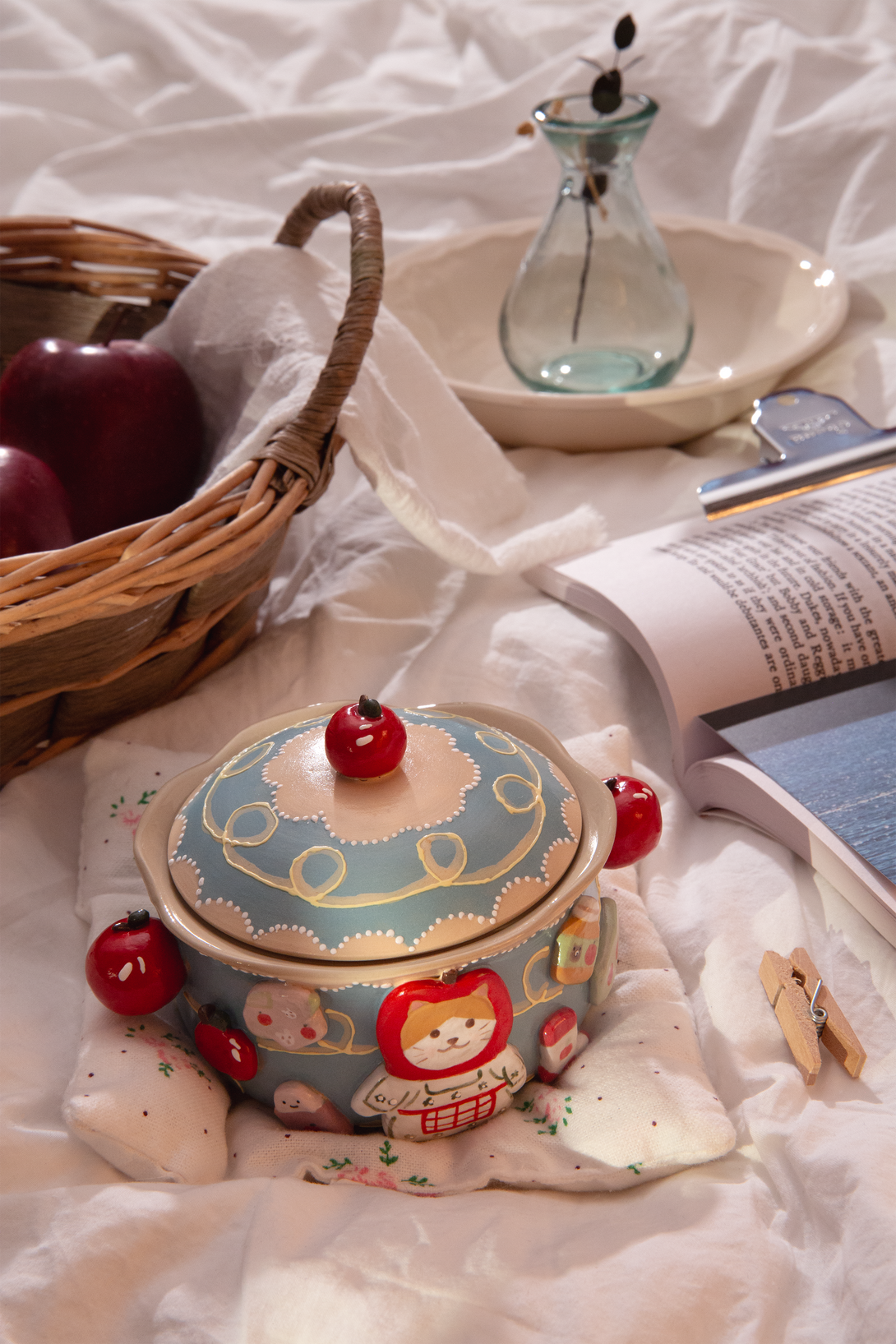 ceramic bowl with lid featuring a cartoon character, surrounded by a basket, vase, and book on a light fabric background.