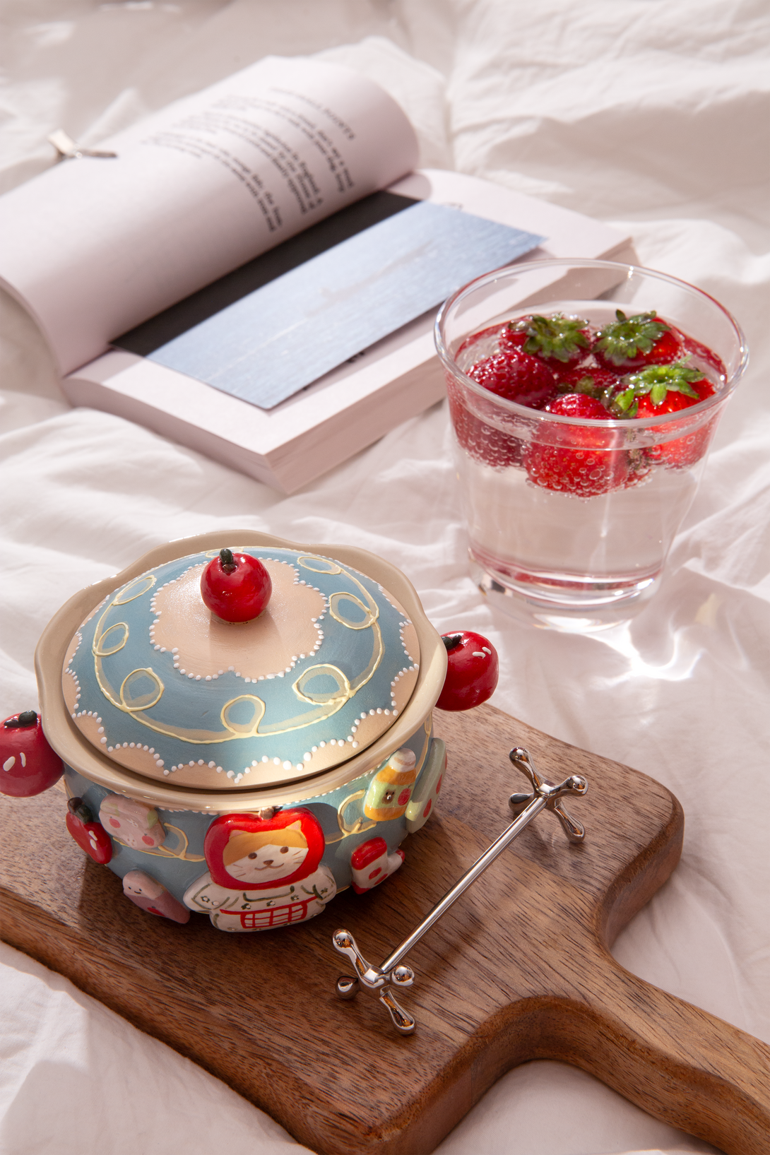 ceramic bowl with a lid, glass of water with strawberries, and open book on a light surface