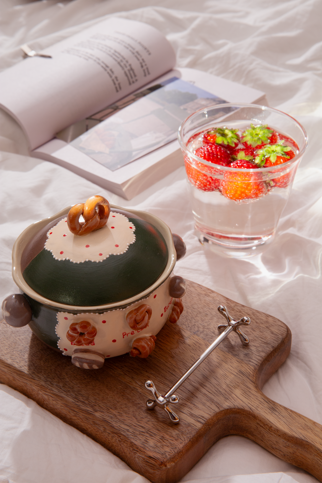 Ceramic bowl with a lid on a wooden board, next to a glass with fruit salad, on a white surface with an open book.