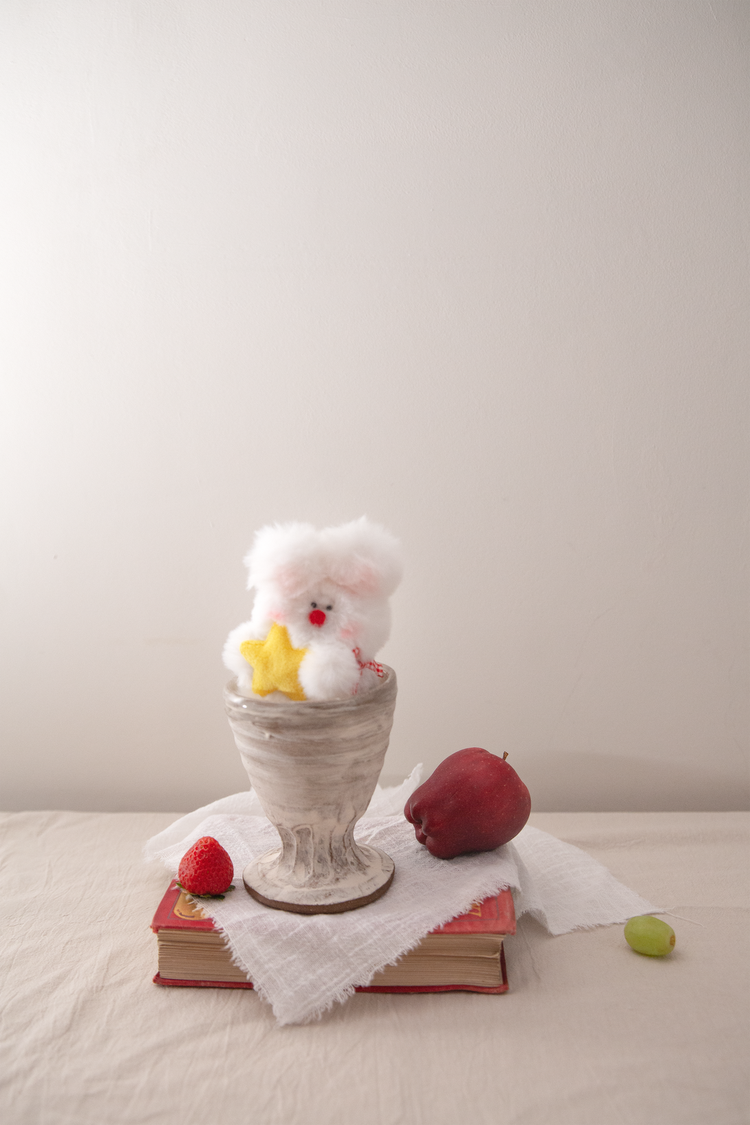 Small plush bag charm on a decorative stand with books and fruits on a light surface.