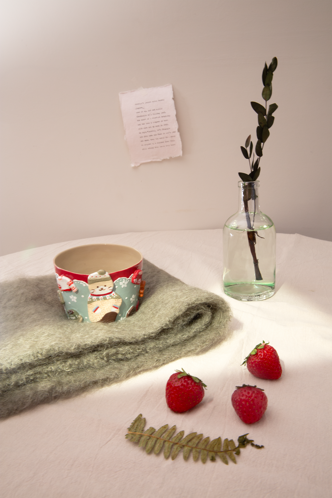 Ceramic cup with Christmas theme design, green towel, strawberries, and a small plant in a glass bottle on a light surface.