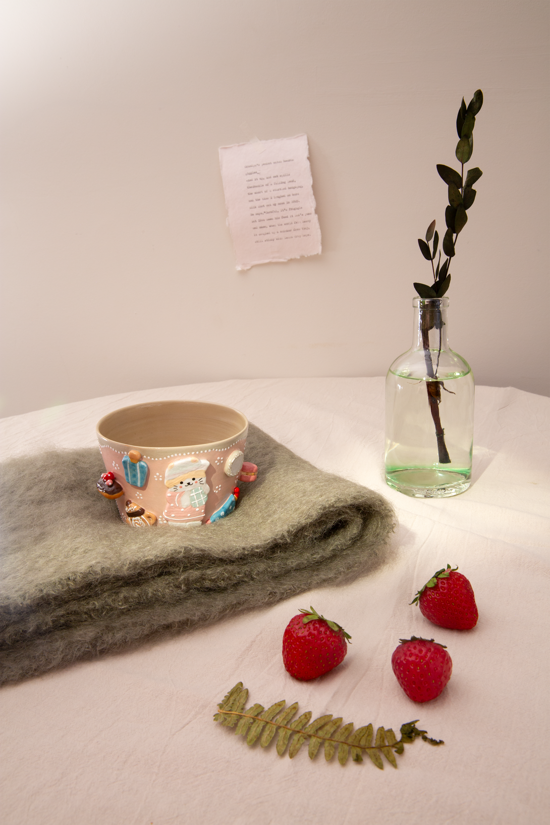 Cup with cartoon characters, folded blanket, strawberries, and a vase on a light surface.