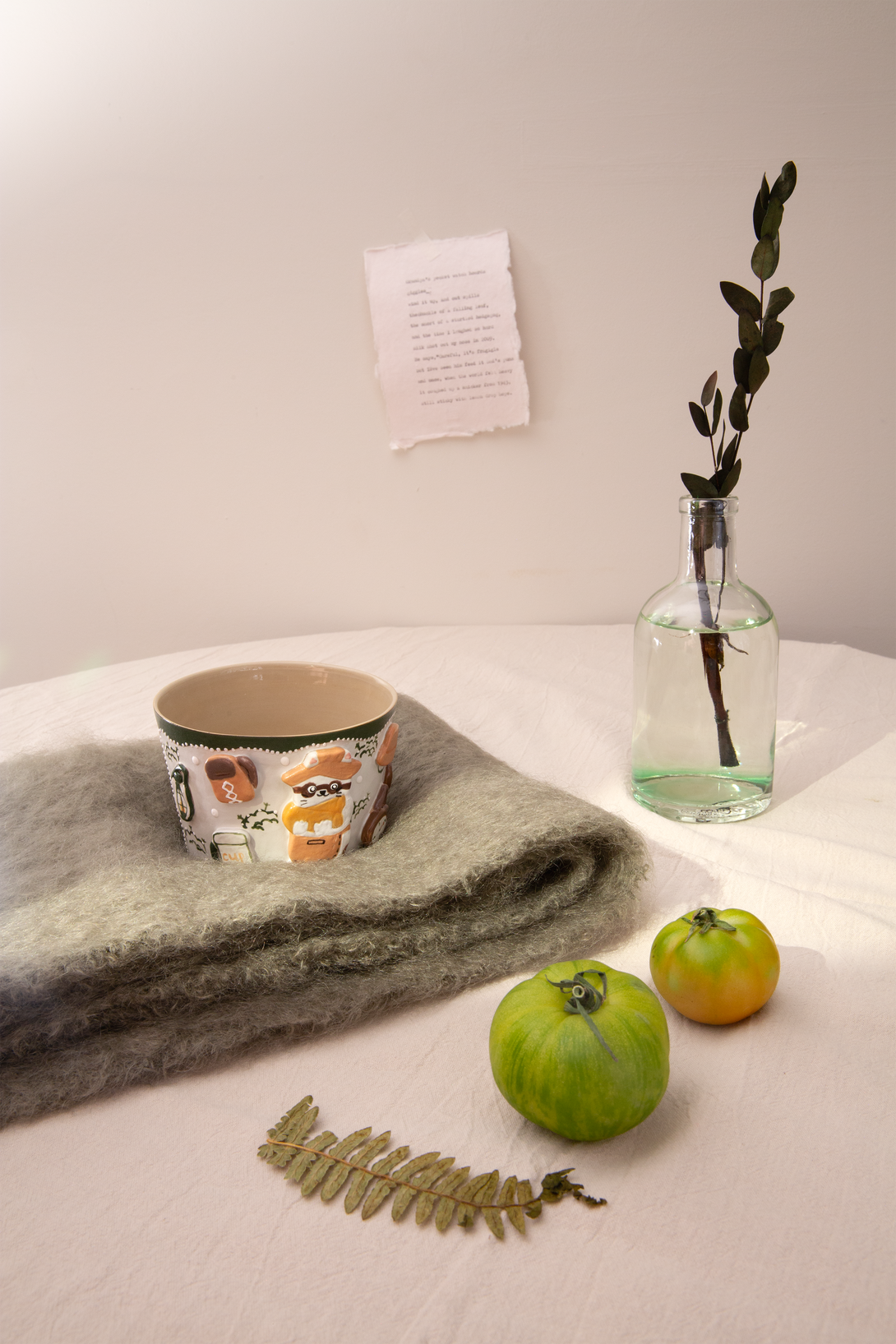 Ceramic mug with animal design, folded fabric, bottle with plant, and fruits on a light surface.