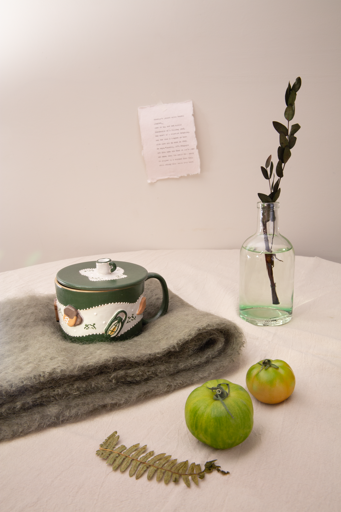 Green mug with 3D design on a blanket, next to a vase with a plant and fruits on a light surface.