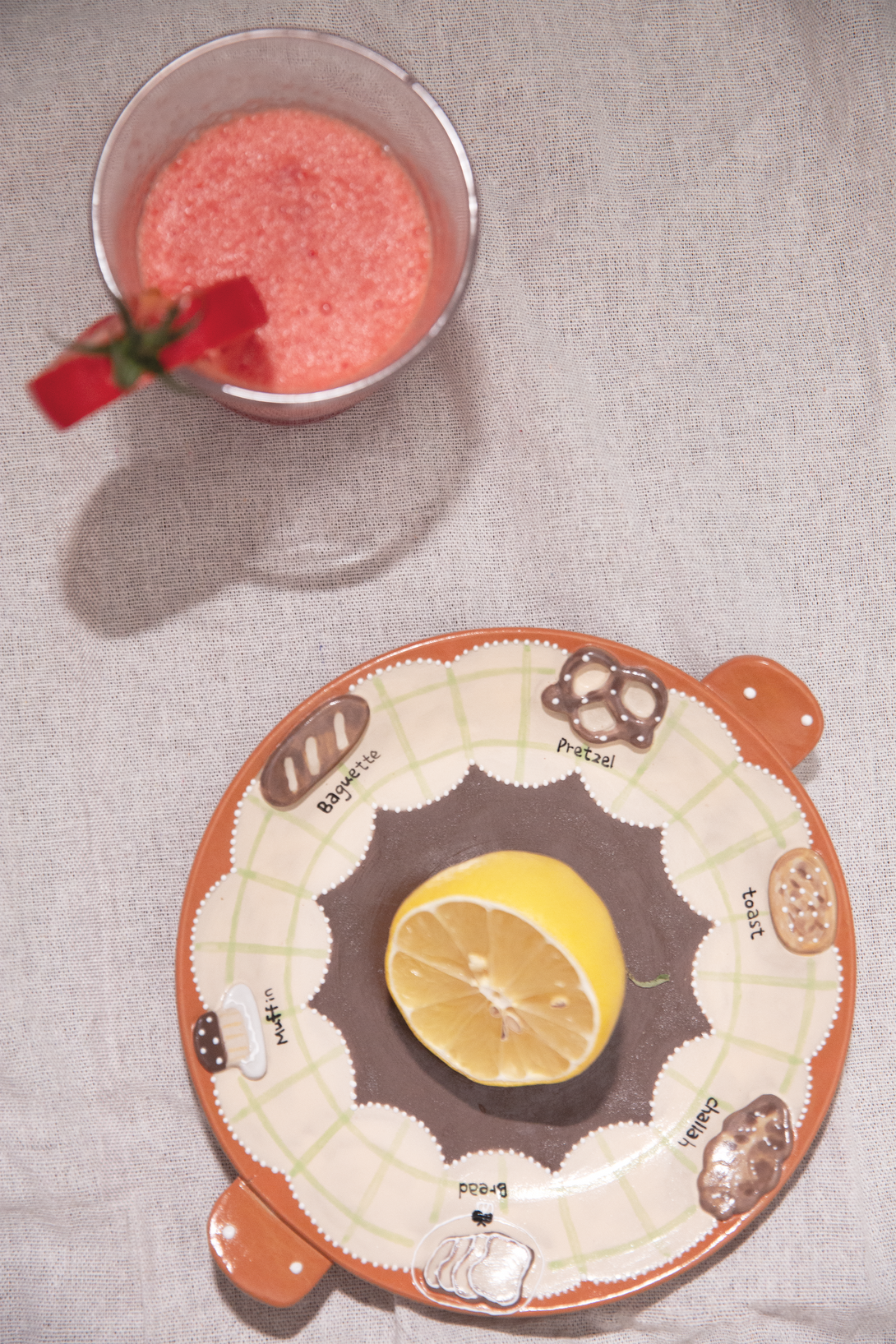Round ceramic plate with a lemon slice on a textured surface next to a cup of pink drink.