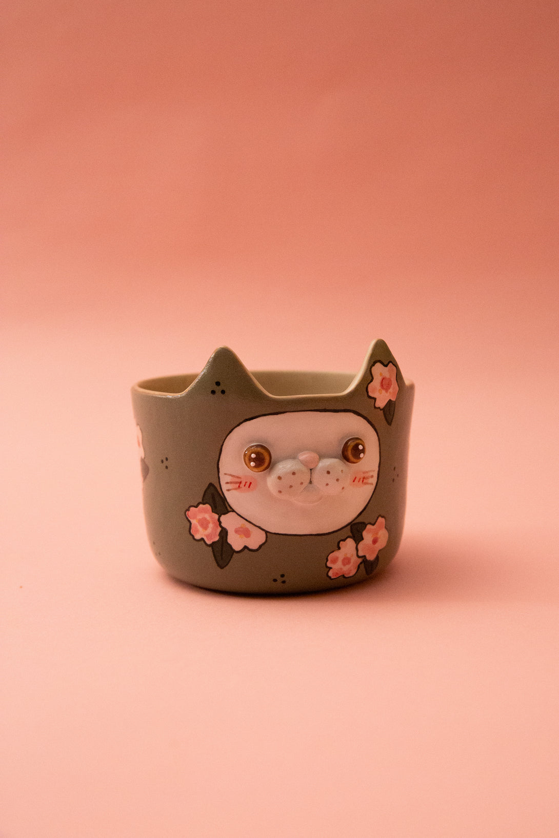 Ceramic cat-shaped mug with face and flowers on a pink background